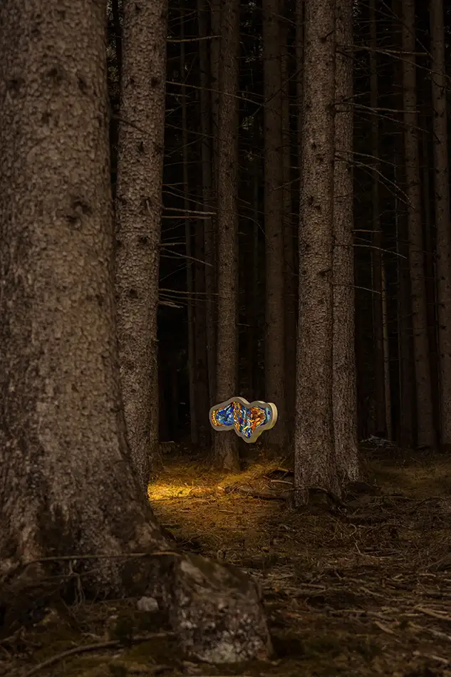 Seitensansicht einer frei hängenden und beleuchteten Bleiverglasung mit dem Namen Amoeba die in einem Wald hängt