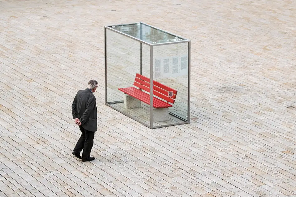 Person vor Kunstinstallation im öffentlichen Raum die eine eingeglaste Parkbank zeigt mit dem Titel Nehmen Sie Platz!