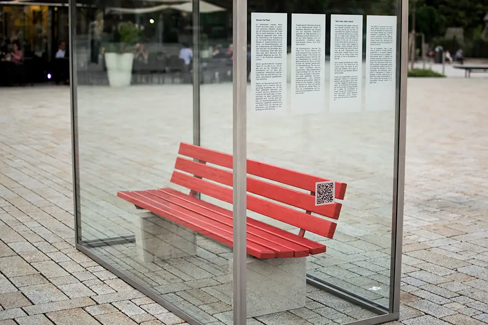 Beschreibungstext auf Glasscheibe von Kunstinstallation im öffentlichen Raum die eine eingeglaste Parkbank zeigt mit dem Titel Nehmen Sie Platz!