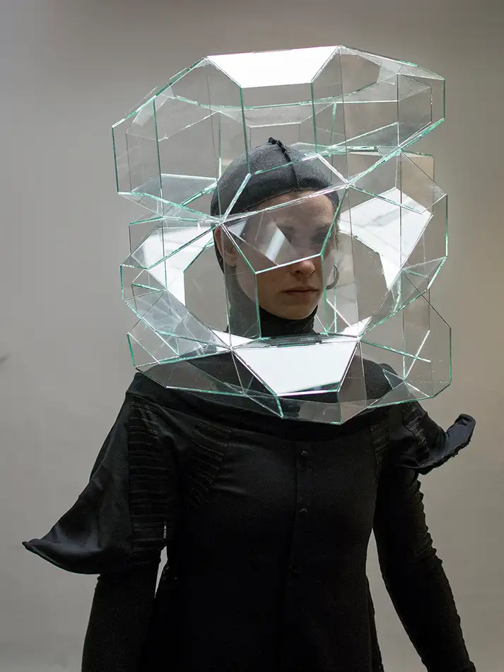 A geometric glass sculpture made of colourless glass that is placed over the head of a person wearing a fashionably simple black dress