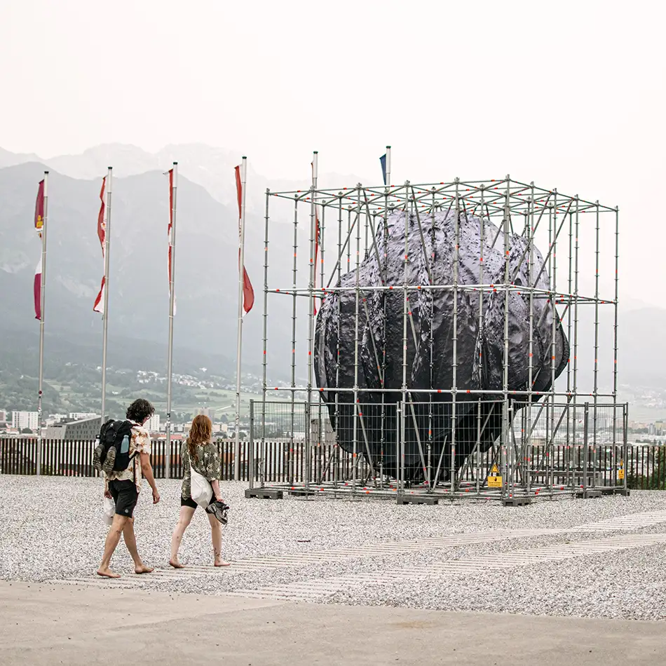 We Are the Asteroid - Kunst im öffentlichen Raum - Ein Asteroid in einer Baugerüststruktur mit Personen und Fahnen im Hintergrund